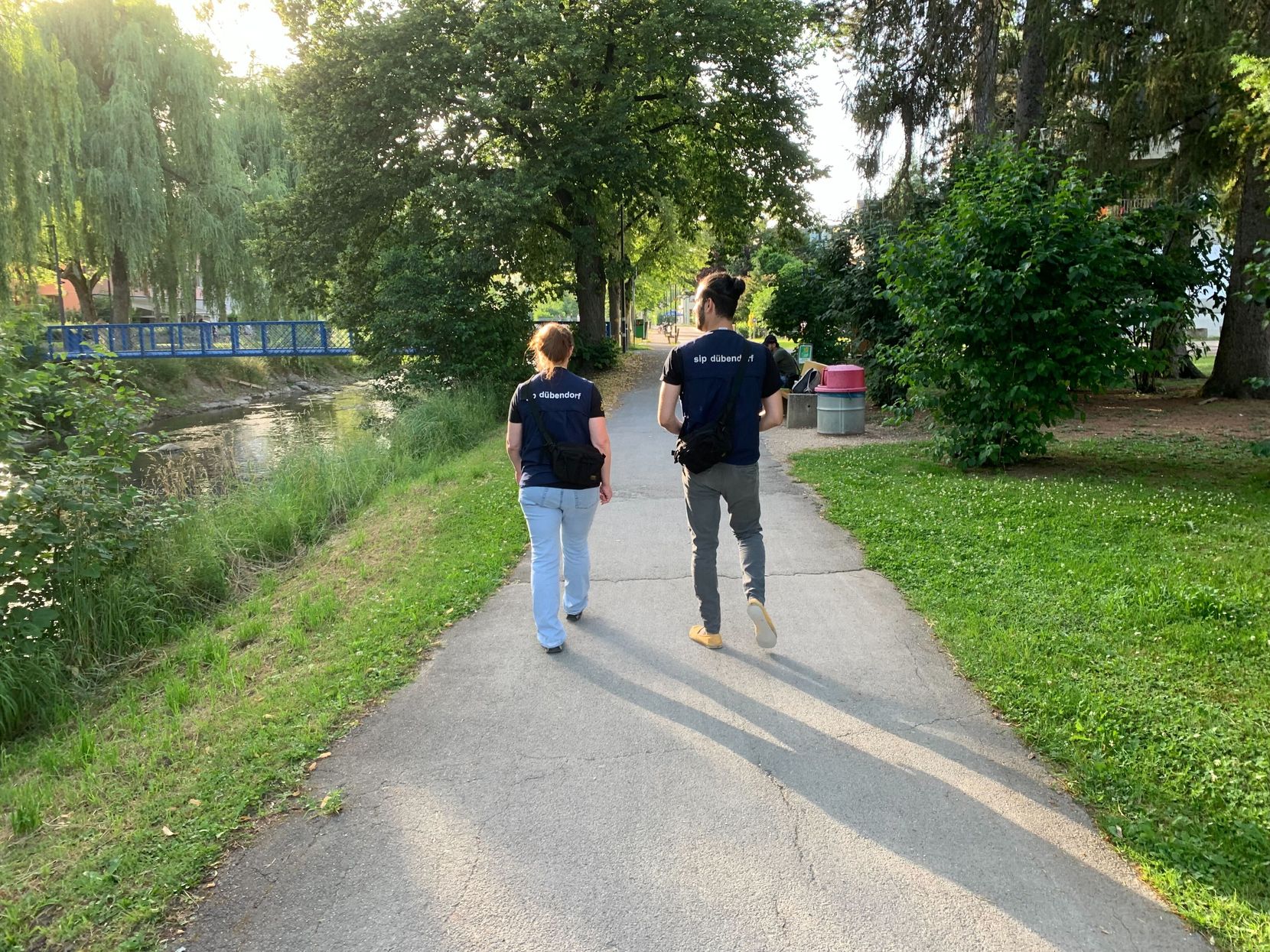 Zwei Personen spazieren auf einem Weg entlang einem Fluss; man sieht sie von hinten.